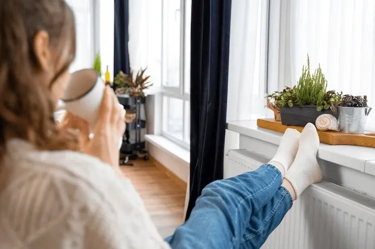 Mujer toma un café mientras apoya los pies en un radiador blanco en su casa.