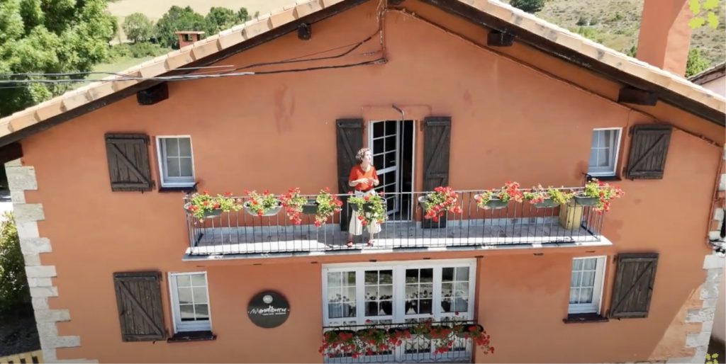 Una vista frontal de una casa rural. Una mujer se ve asomada a un gran balcón de la fachada principal.